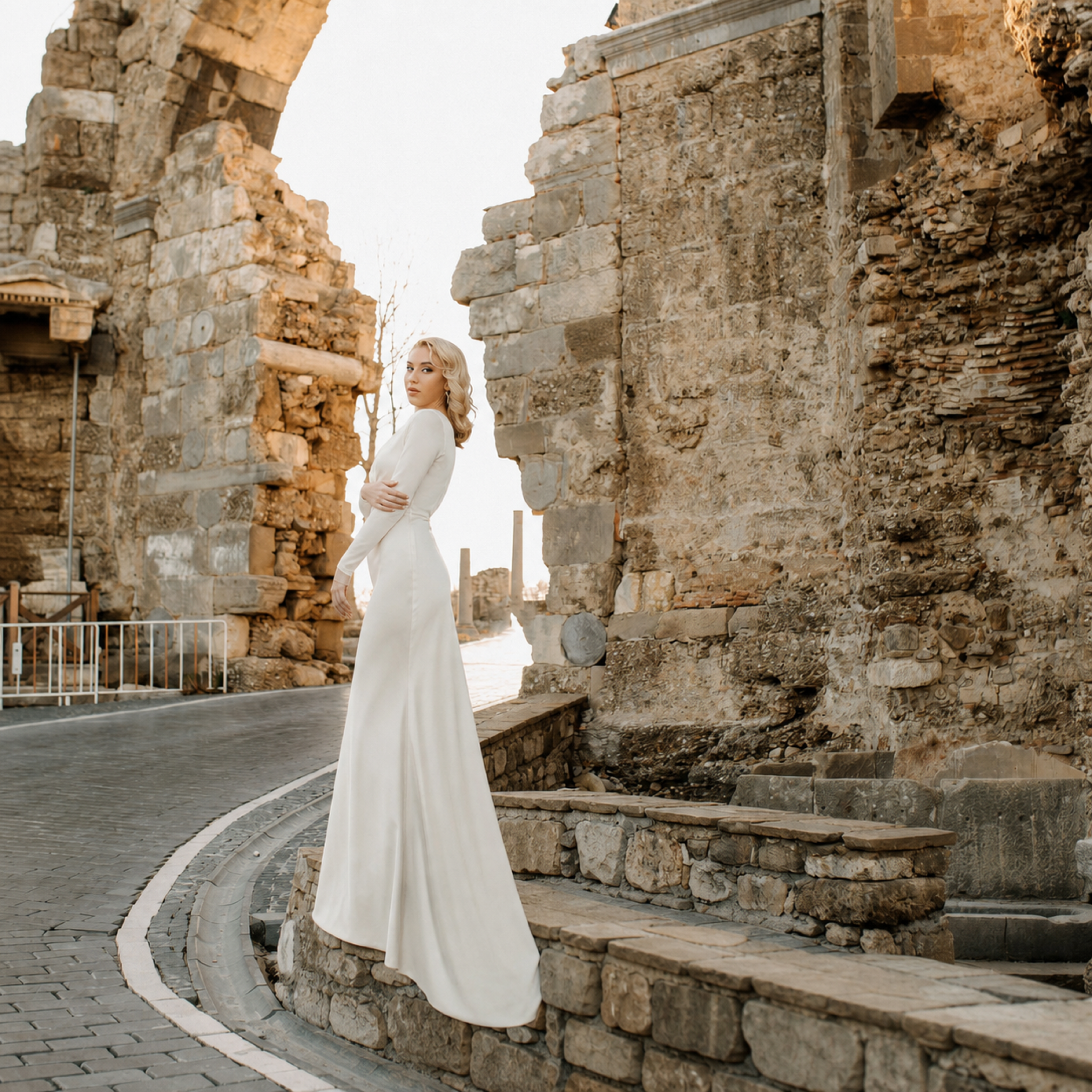 Model in an ivory long-sleeve, boat-neck floor-length Scarlett dress beneath stone arches.